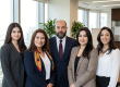 Five colleagues in business attire posing for a group photo in a bright office.