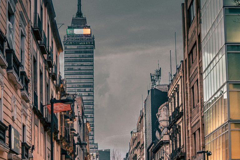 City street viewed from ground level, with historic buildings on either side and a tall modern tower in the distance against a cloudy sky.