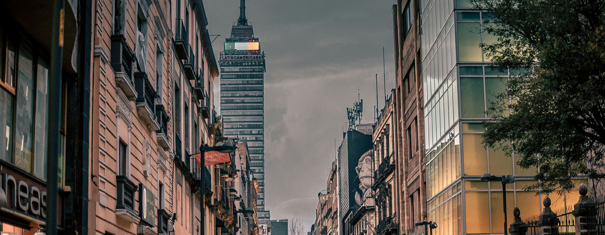 City street viewed from ground level, with historic buildings on either side and a tall modern tower in the distance against a cloudy sky.