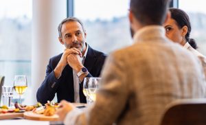 A middle-aged man in a dark blazer sits at a dining table with his hands clasped, listening thoughtfully during a business meal, with wine glasses and plates of food in front of him.