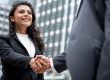 Two professionals shake hands in front of a city office building, signaling a business agreement or greeting.