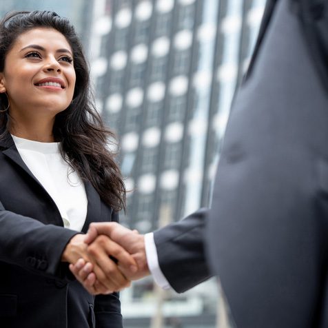 Two professionals shake hands in front of a city office building, signaling a business agreement or greeting.