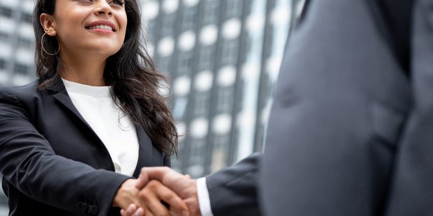 Two professionals shake hands in front of a city office building, signaling a business agreement or greeting.