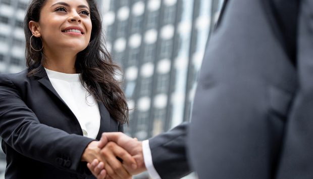 Two professionals shake hands in front of a city office building, signaling a business agreement or greeting.