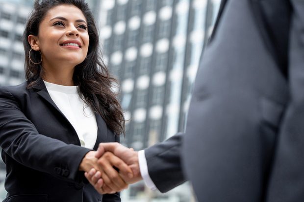 Two professionals shake hands in front of a city office building, signaling a business agreement or greeting.
