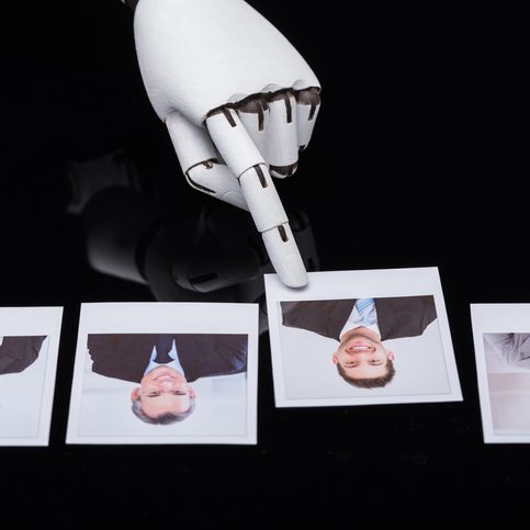 Robotic hand with extended finger points at a row of four Polaroid photos of businesspeople on a dark surface.
