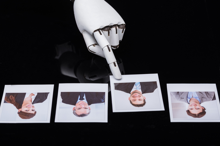 Robotic hand with extended finger points at a row of four Polaroid photos of businesspeople on a dark surface.